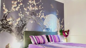 Un dormitorio con cama, mesita de noche, un mural decorativo y un jarrón con flores.