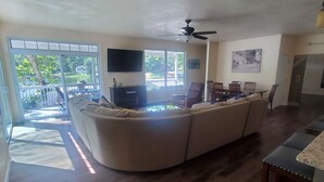 Una sala de estar con un amplio sofá seccional en forma de L, un televisor de pantalla plana montado en la pared y un ventilador de techo. Hay un comedor con una mesa y sillas, y vistas a un lago a través de amplias ventanas.