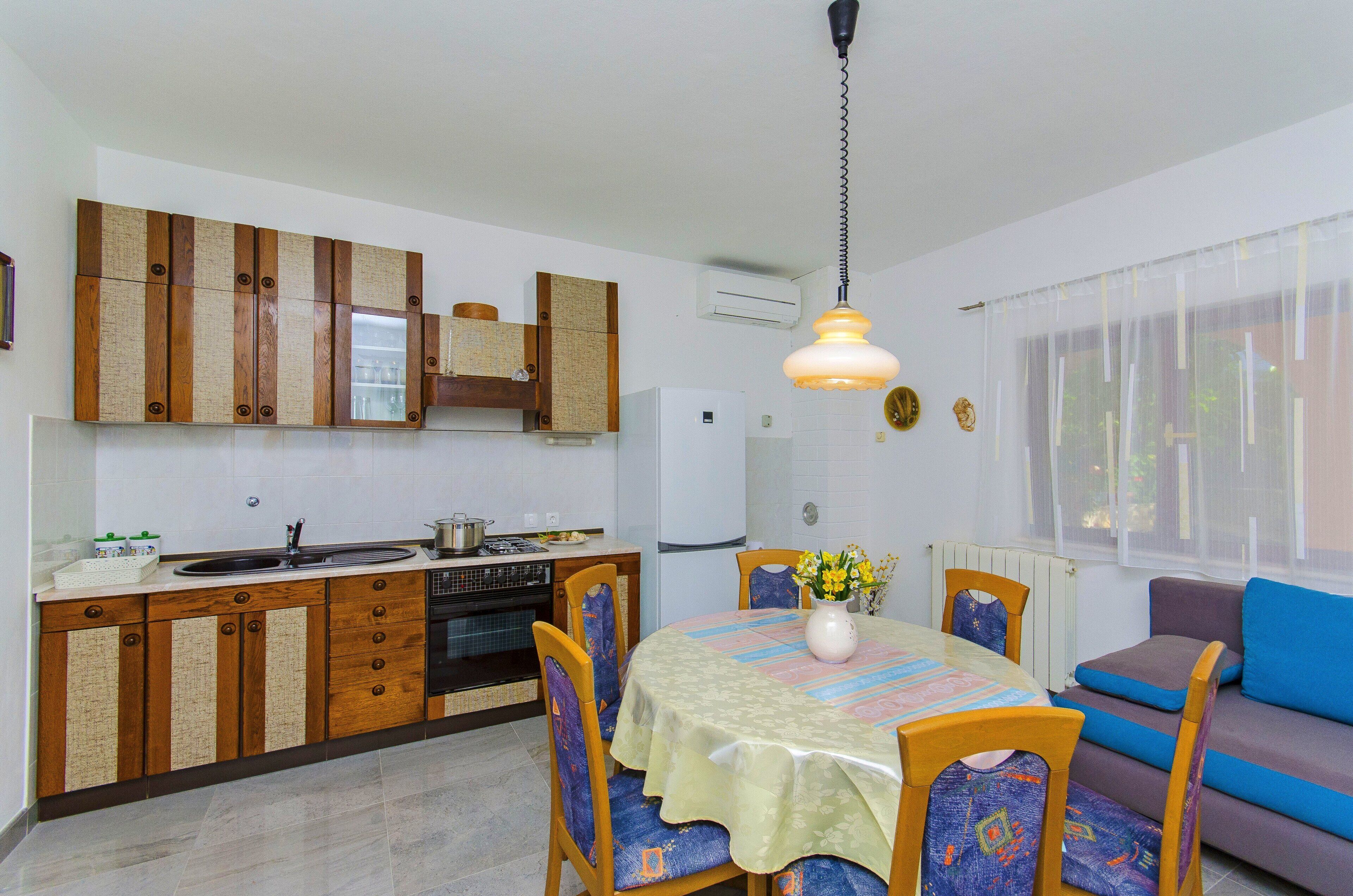 A compact kitchen with wooden cabinets, a dining table with chairs, a sofa, and a hanging light fixture.