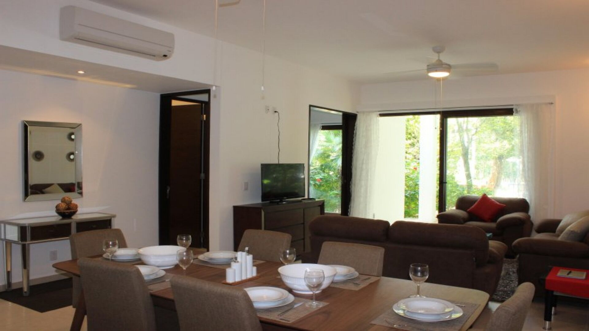 A modern living room with a dining area, a flat-screen TV, and a large window with a view of trees.