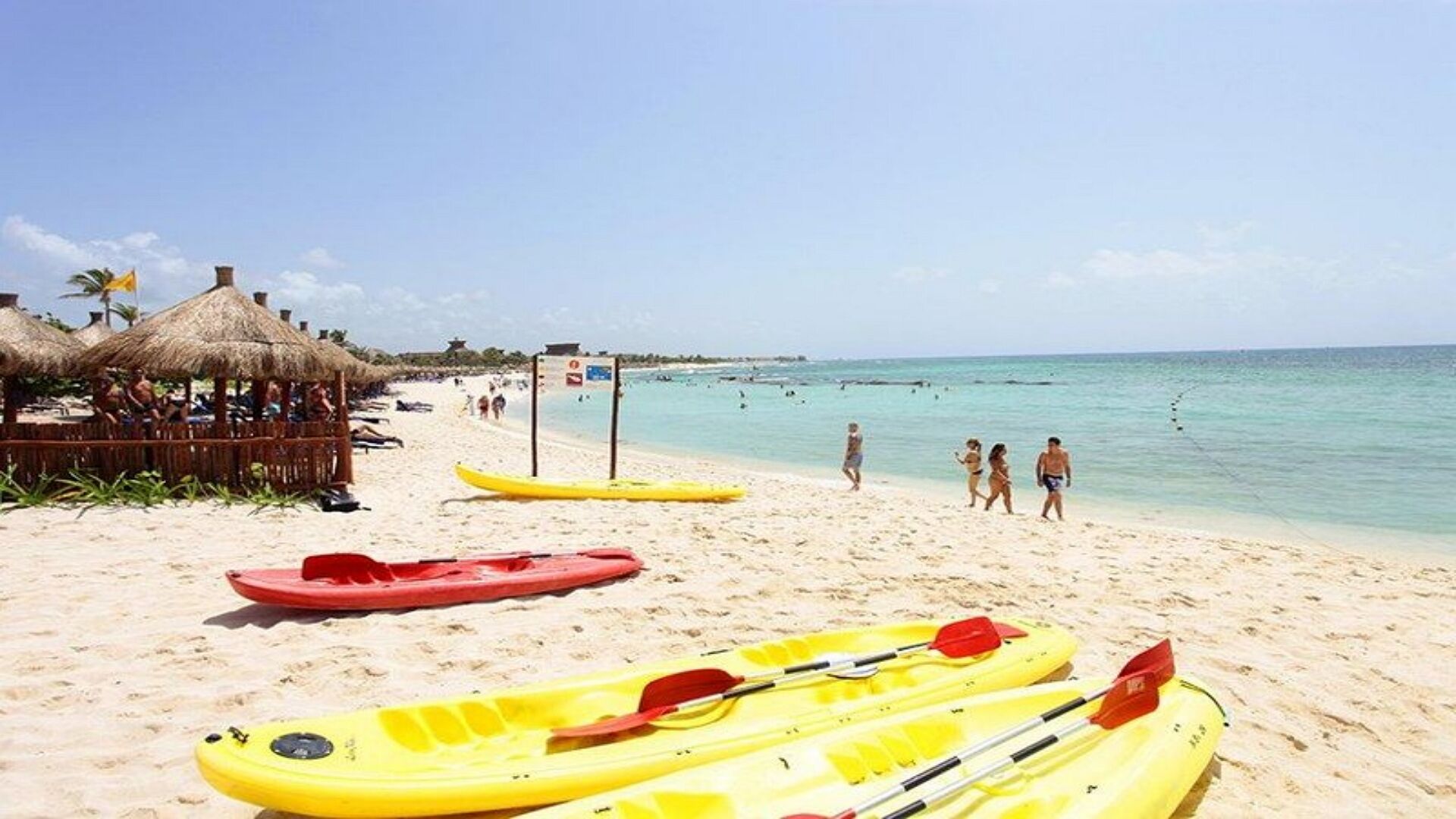 Una playa con kayaks amarillos, una cabaña con techo de paja y gente caminando por la orilla.