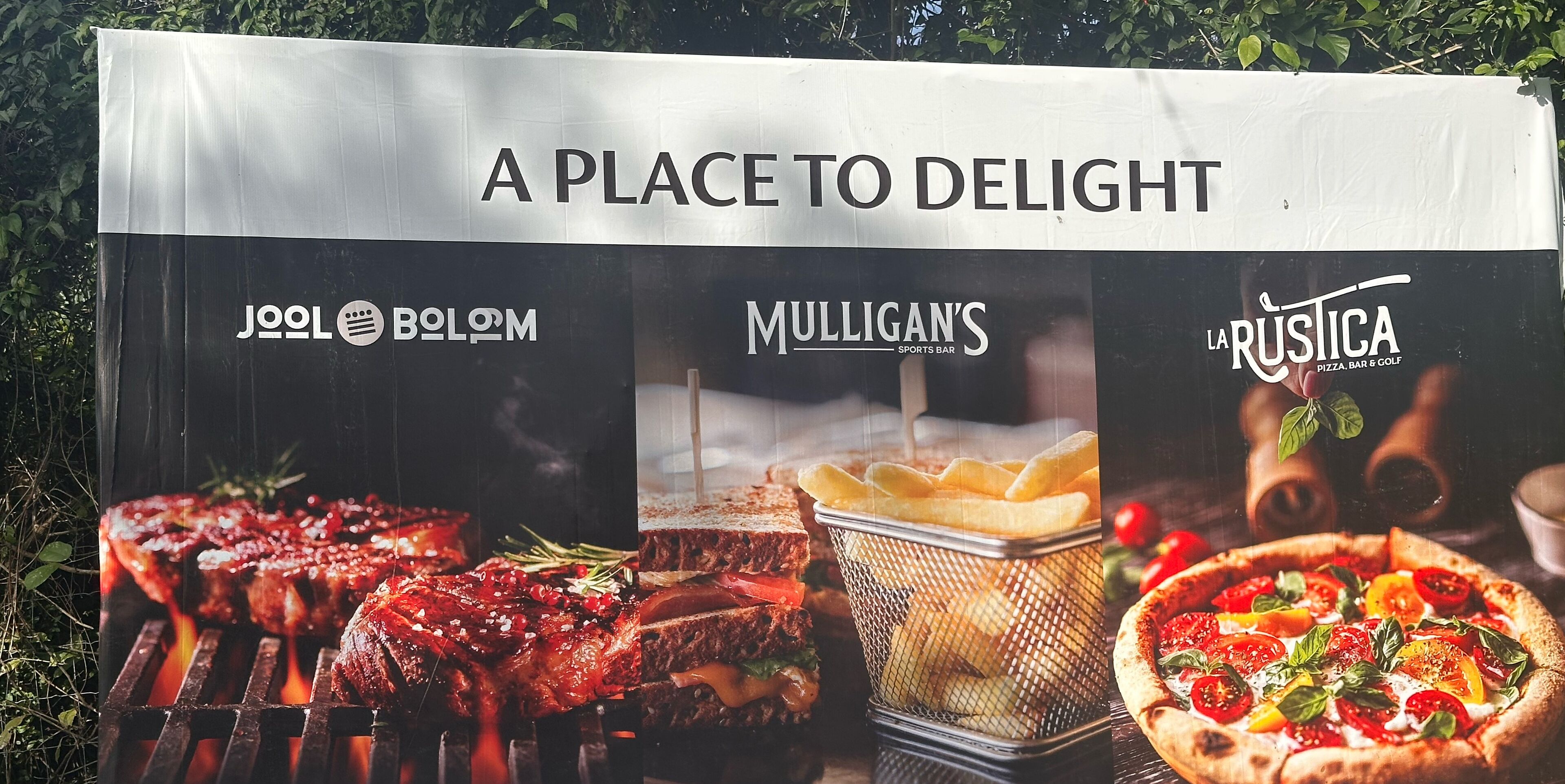A signboard with the text "A PLACE TO DELIGHT" and logos of Jool, Bolom, Mulligan's, and La Rustica.