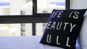 A pillow with the text "Life is Beautiful" on a bed with a view of buildings through a window. - Diamond Head and Ocean View Waikiki Sunset! (WS06) (Honolulu)
