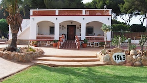 A white building with a red-tiled roof, a staircase leading to a wooden door, and a stone wall with a rock labeled "1963". - See view villa with pool and big garden. (MORAIRA, (ALICANTE) ESPAÑA)
