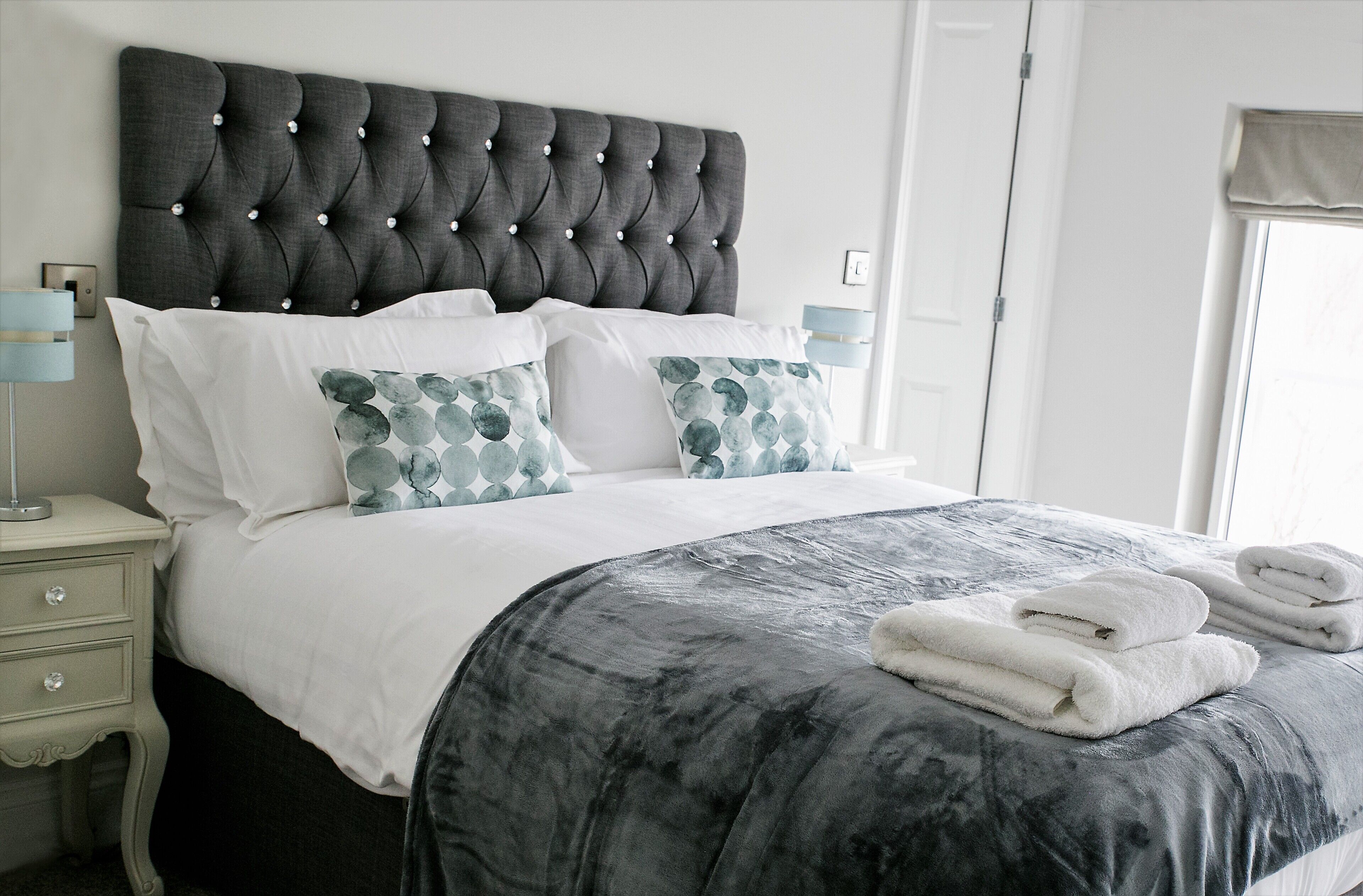 Un lit bien fait, avec une tête de lit capitonnée, une literie blanche et un plaid à motifs gris et blanc.