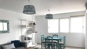 Ein modernes Wohnzimmer mit einem grauen Sofa, einem Holztisch, einem weißen Bücherregal, blaugrünen Esszimmerstühlen und einer blauen Pendelleuchte.