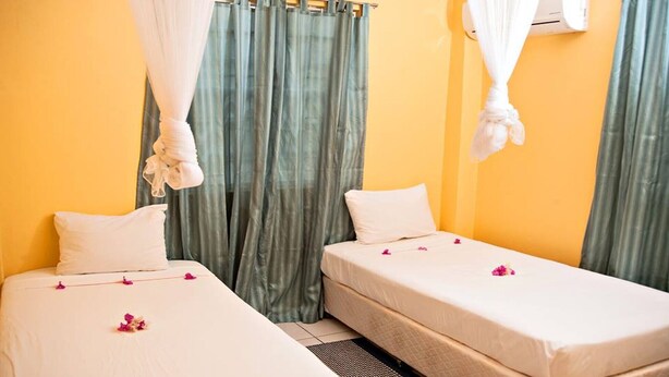 A room with two single beds, each with a white bedspread and pillows, and a small bedside table. The room has a yellow wall, a green curtain, and a white air conditioning unit.