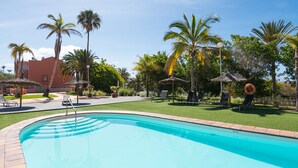 Una piscina con sillones de descanso y sombrillas, rodeada de palmeras y un edificio al fondo.