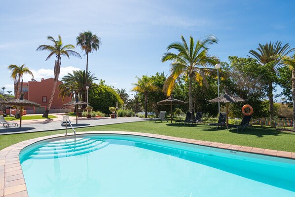A swimming pool with lounge chairs and umbrellas, surrounded by palm trees and a building in the background.