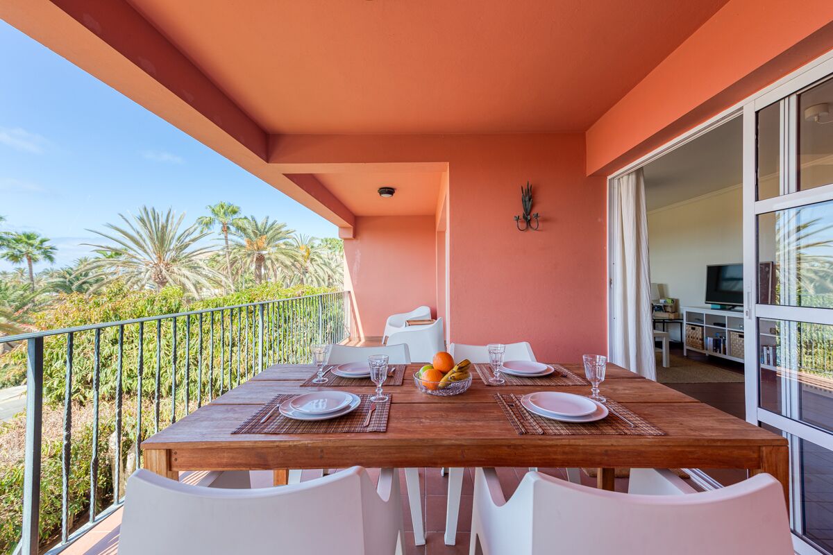 A balcony with a wooden table set for dining, white chairs, and a view of palm trees.