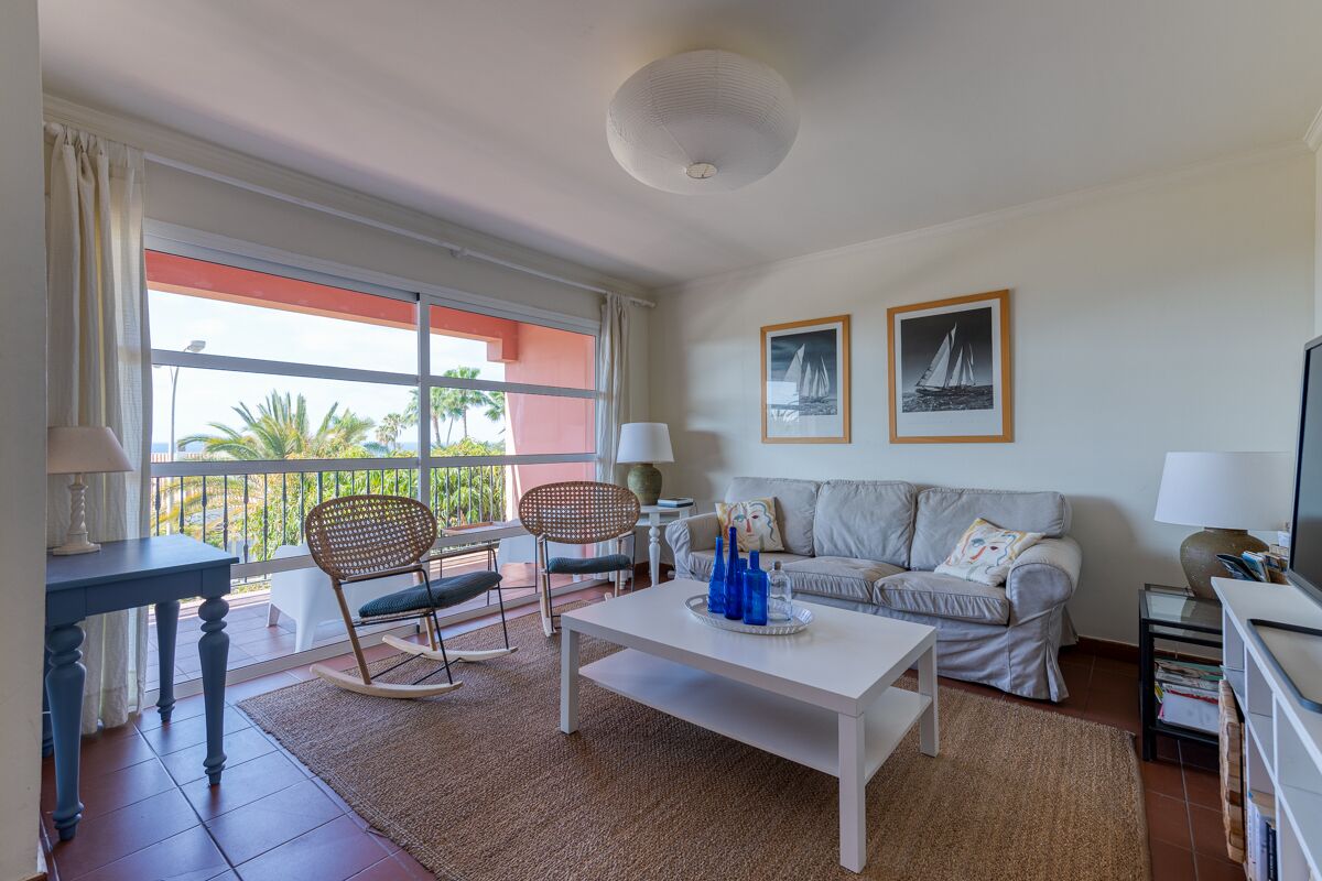 A living room with a white sofa, a rocking chair, a coffee table, and a TV stand. There are two framed pictures on the wall and a window with a view of palm trees.