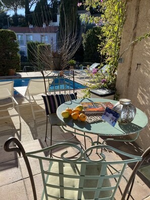 Une terrasse extérieure ensoleillée avec une piscine, des chaises longues et une table où sont disposés des fruits et un livre.