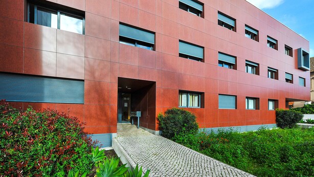 Een modern gebouw met een rode gevel, meerdere ramen en een verharde entree met beplanting.