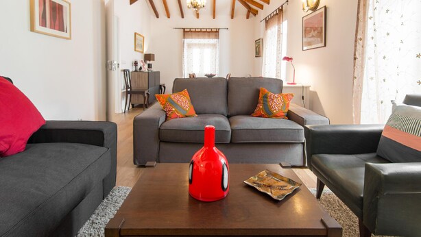 Un salon avec un canapé, une table basse sur laquelle se trouve un objet rouge, et un coin repas à l’arrière-plan.