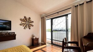 Ein Hotelzimmer mit Bett, Fernseher, Wanddeko, einem kleinen Holzschrank, einem Sessel und einem Balkon mit Vorhängen.