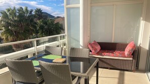 En balkon med fletmøbler, et glasbord og udsigt til palmer og bakker.