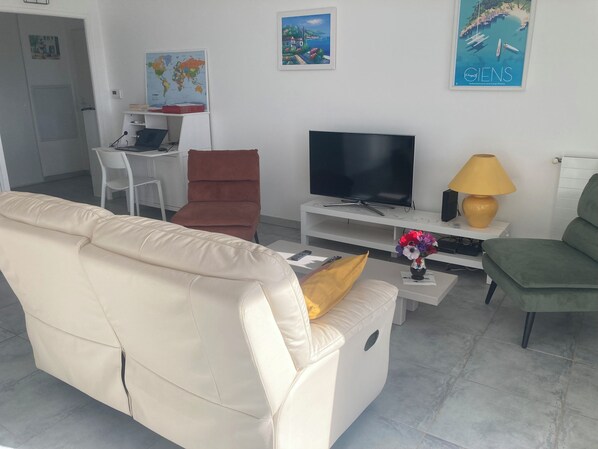 A living room with a beige sofa, a brown chair, a TV on a white stand, a lamp, and a small table with a vase.