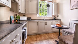 A kitchen with light-colored cabinets, a window with a view, a sink, a stove, and a dining area with a table and chairs. - The Clock House at Heligan Gardens (Pentewan)