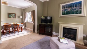 A living room with a fireplace, a television, a dining area with chairs and a table, and a painting on the wall. - The Clock House at Heligan Gardens (Pentewan)
