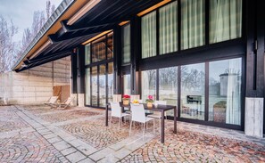A covered outdoor seating area with a table and chairs, large windows, and a view of a body of water.