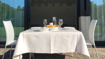 Un espace de restauration en plein air avec une table dressée pour deux personnes, agrémentée d’une nappe blanche, de deux verres à vin, d’une bouteille de vin et d’un panier de pain.
