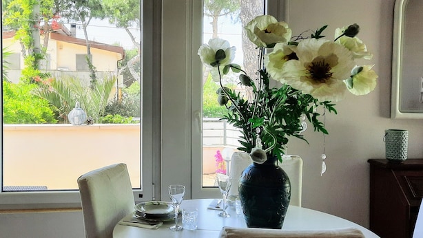Un tavolo da pranzo con un vaso di fiori, una sedia e una finestra che si affaccia su vegetazione ed edifici.