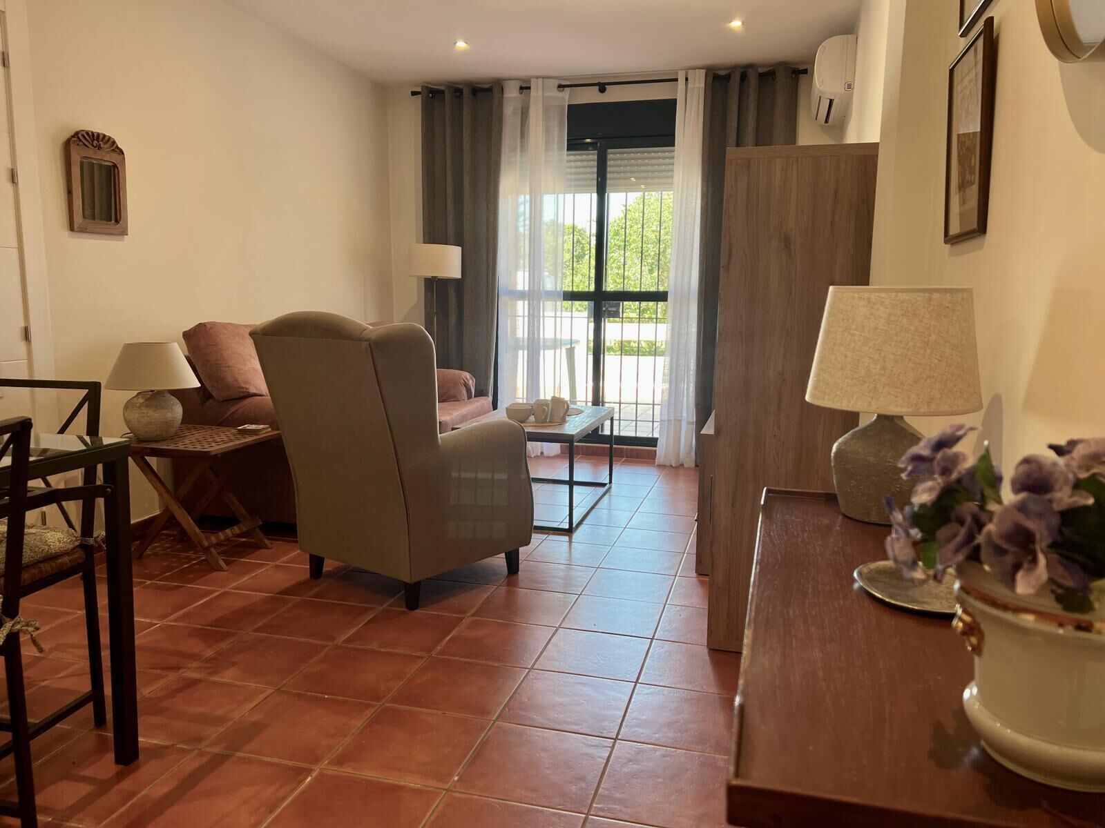 A living room with a sofa, armchair, coffee table, and a dining table. There is a window with curtains, a door, and a wall-mounted air conditioning unit.