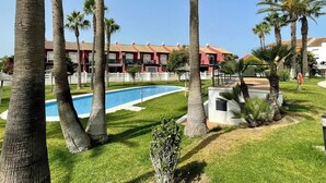 Une piscine entourée de palmiers, un bâtiment rouge doté de balcons et un ciel d’un bleu limpide.