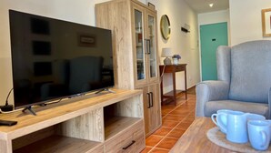 Un salon équipé d’un téléviseur à écran plat, d’un meuble en bois, d’un fauteuil gris et d’une petite table sur laquelle sont posées des tasses bleues.