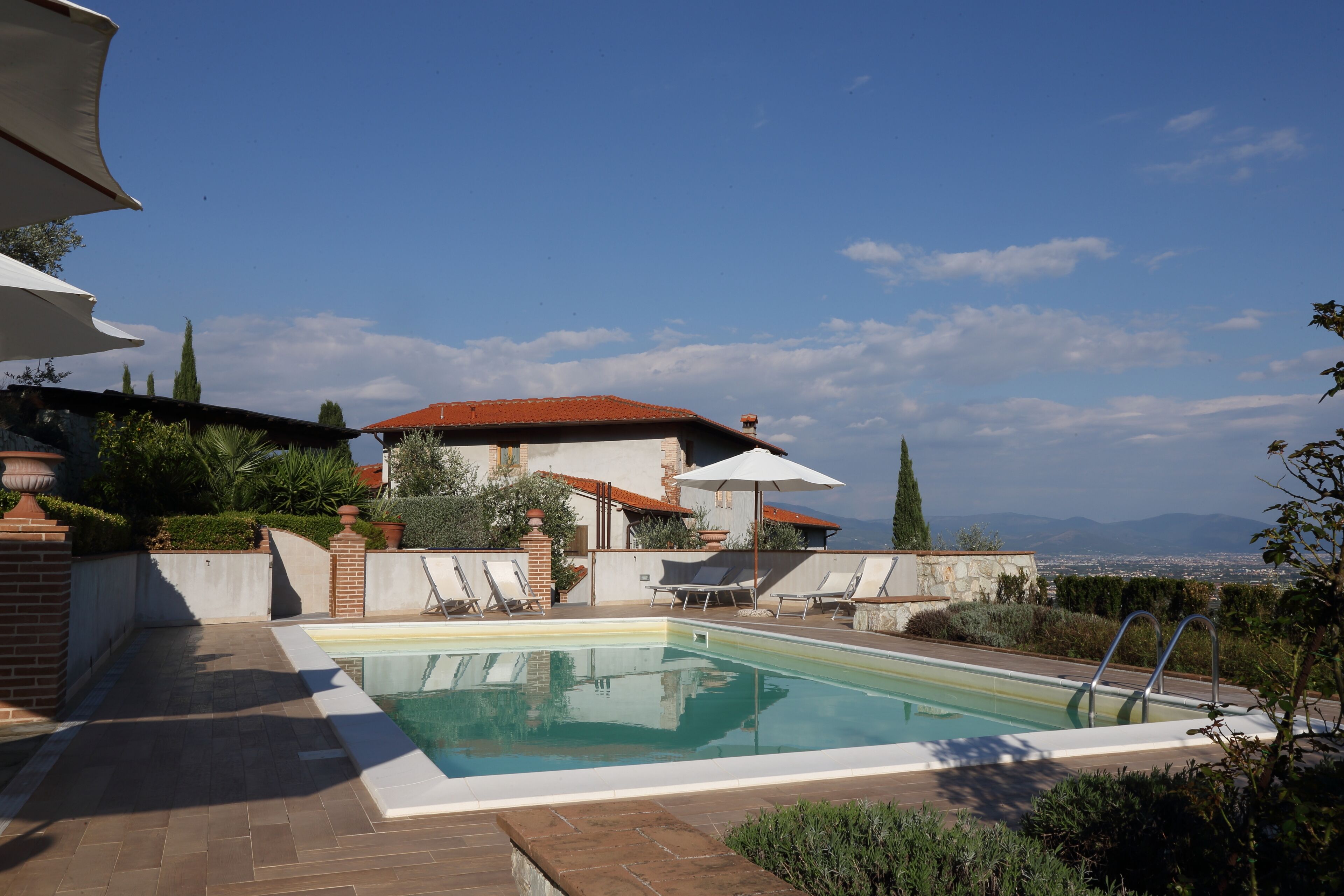 Piscina con sillas de descanso y sombrillas, rodeada por un muro de ladrillo y follaje.