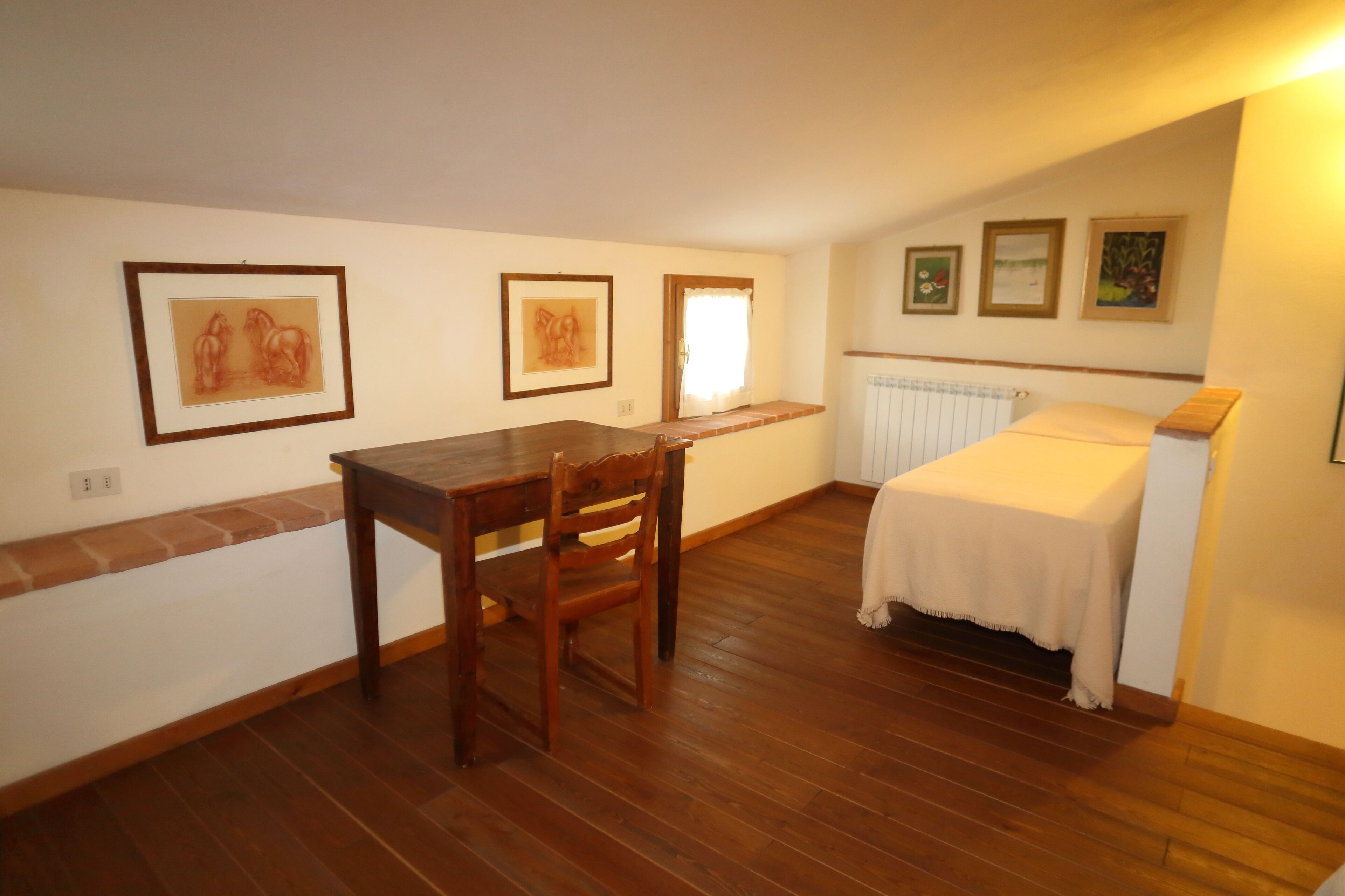 A room with a wooden table and chairs, a bed with a white cover, and three framed pictures on the wall.