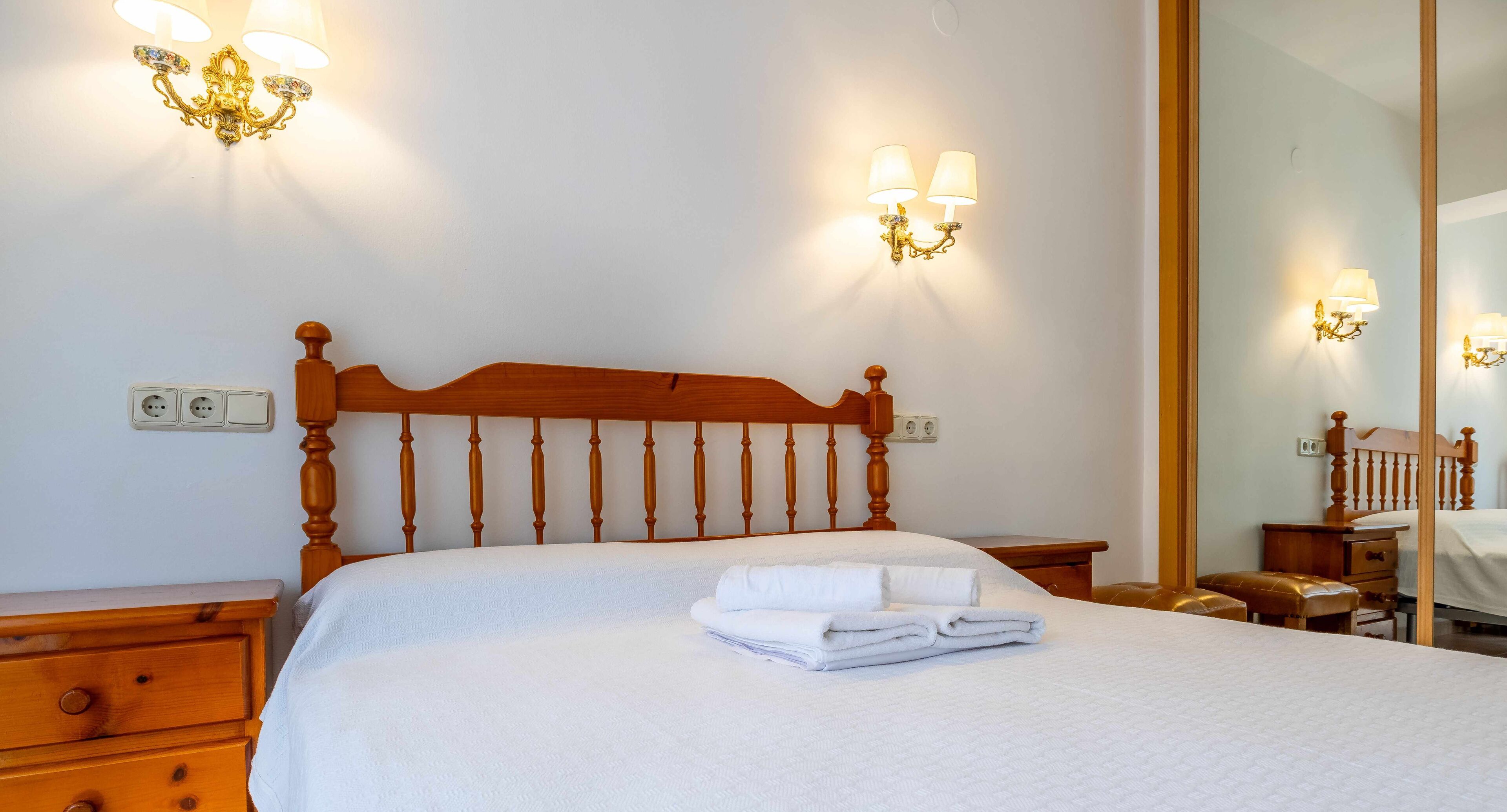 A neatly made bed with a wooden headboard, two wall sconces, a bedside table, and a mirror.