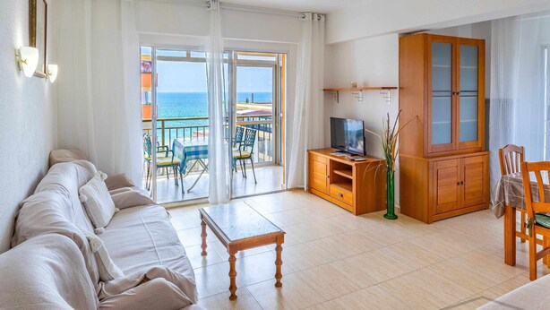 A living room with a sofa, a TV, a wooden cabinet, and a balcony with a view of the sea.