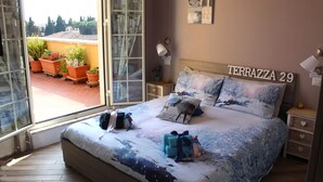 Ein Schlafzimmer mit einem Bett, Kissen, einem Nachttisch, einer Lampe und einem Fenster mit Blumentöpfen draußen.