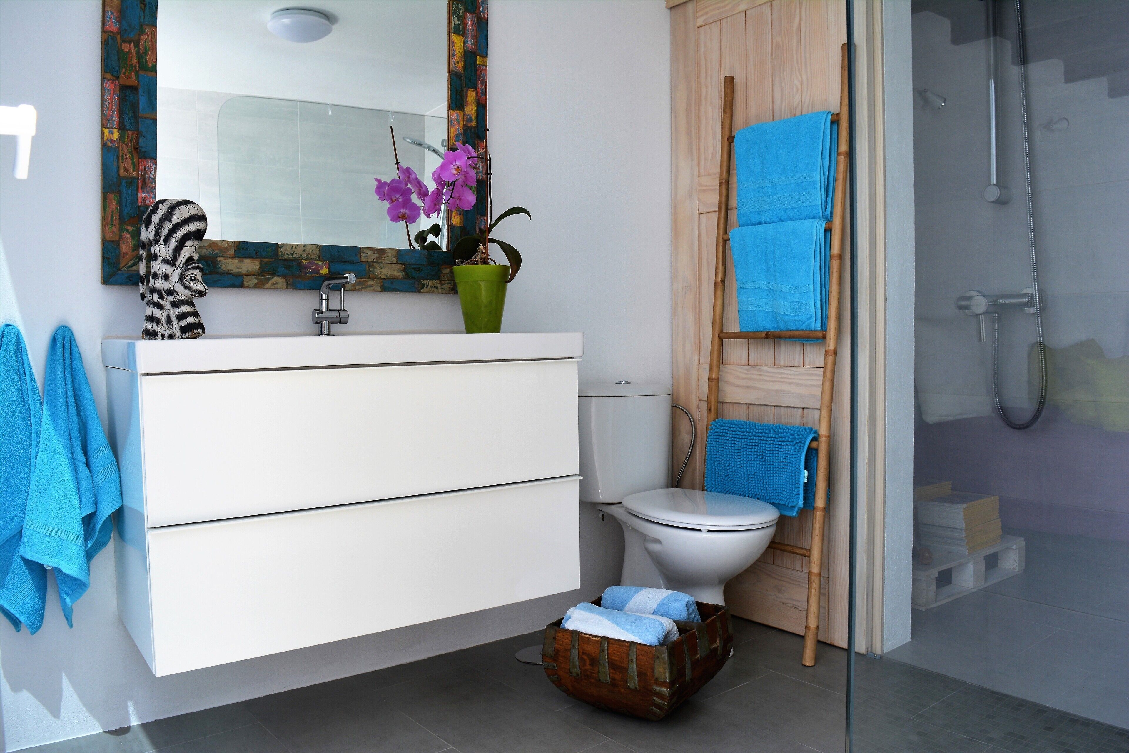 Ein modernes Badezimmer mit einem weißen Waschtisch, einer grünen Vase mit Blumen, einem blauen Handtuch und einer Duschkabine.