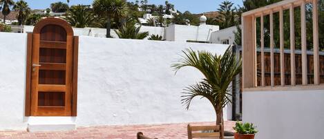 Un patio con una mesa y sillas de madera, una planta en maceta y una pared blanca con una puerta de madera.