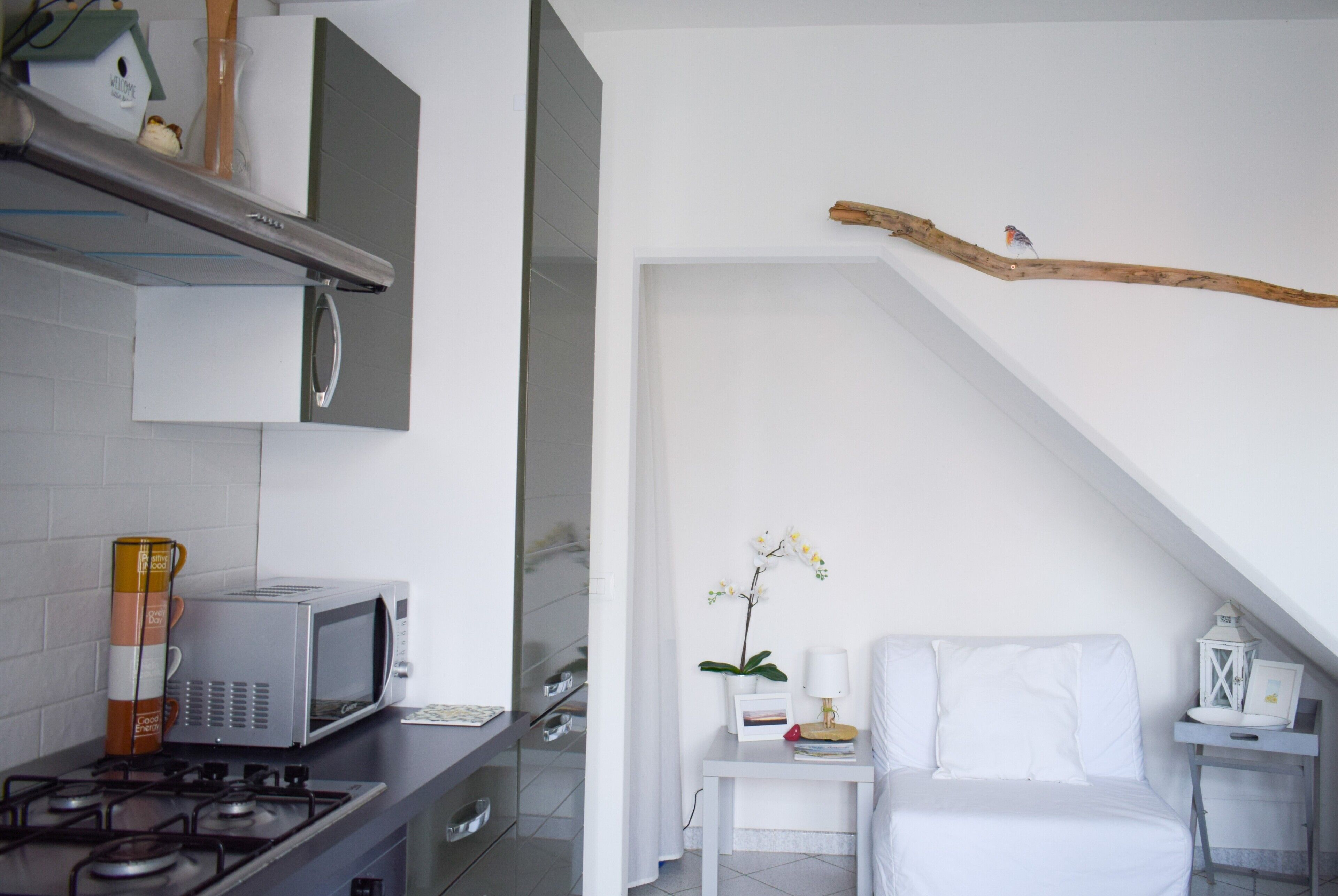 Una cocina moderna con microondas, estufa y armarios. Hay una cama blanca con una mesita de noche y una pequeña mesa con una lámpara y un marco de fotos. Una rama cuelga del techo, y una orquídea está sobre la mesita de noche.
