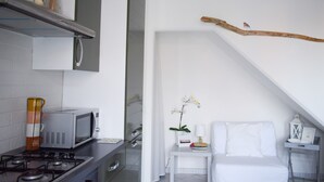 A modern kitchen with a microwave, stove, and cabinets. There is a white bed with a nightstand and a small table with a lamp and a picture frame. A branch hangs from the ceiling, and an orchid is on the nightstand.