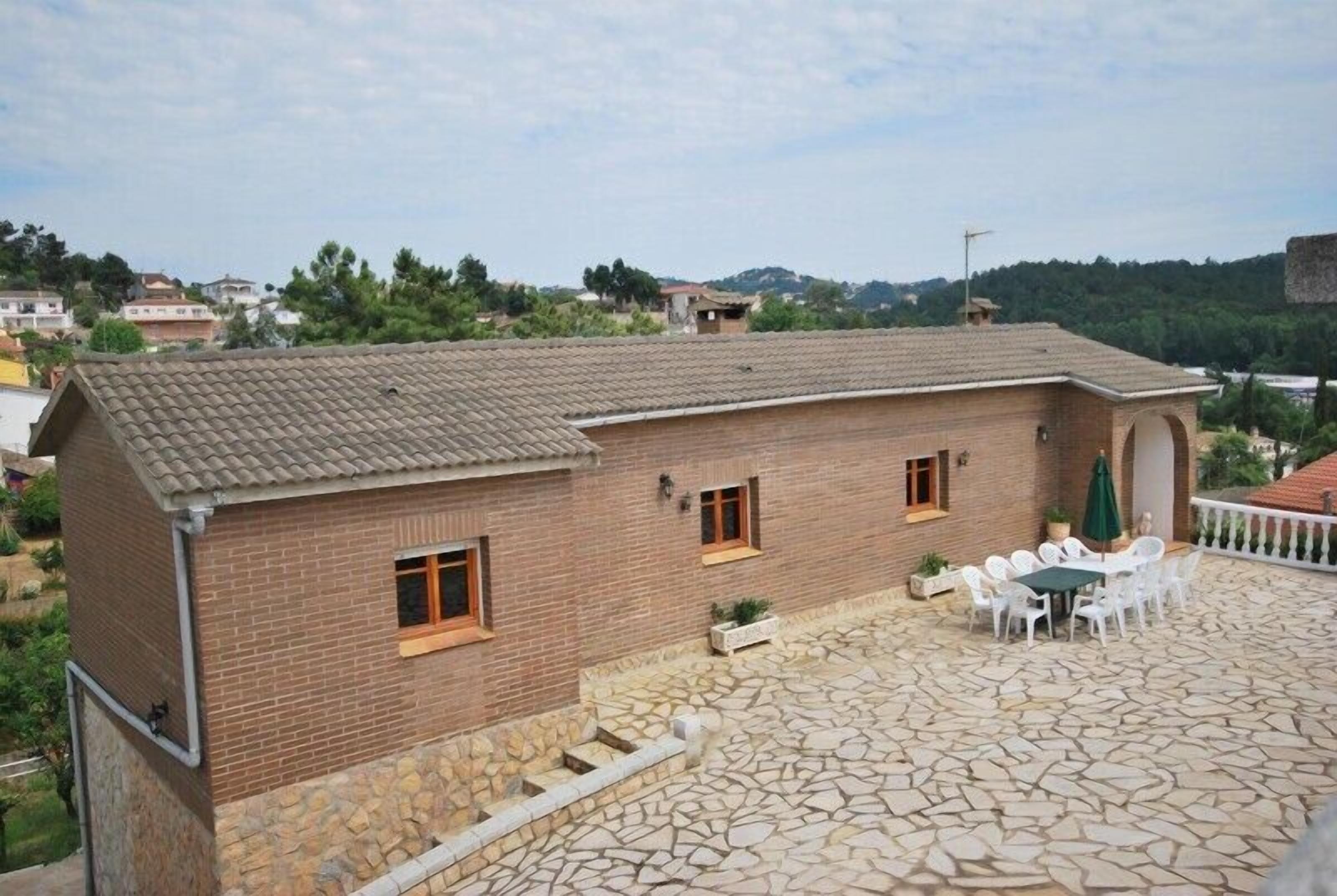 A two-story brick building with a tiled roof, a stone patio with outdoor furniture, and a view of a hilly landscape.