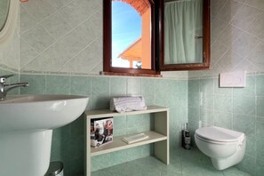 A bathroom with a sink, toilet, shelf with a towel and a magazine, and a window with a view of a building.