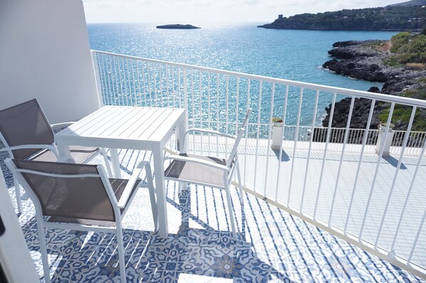 A balcony with a white table and chairs, overlooking a coastal area with rocky terrain and a view of the sea.