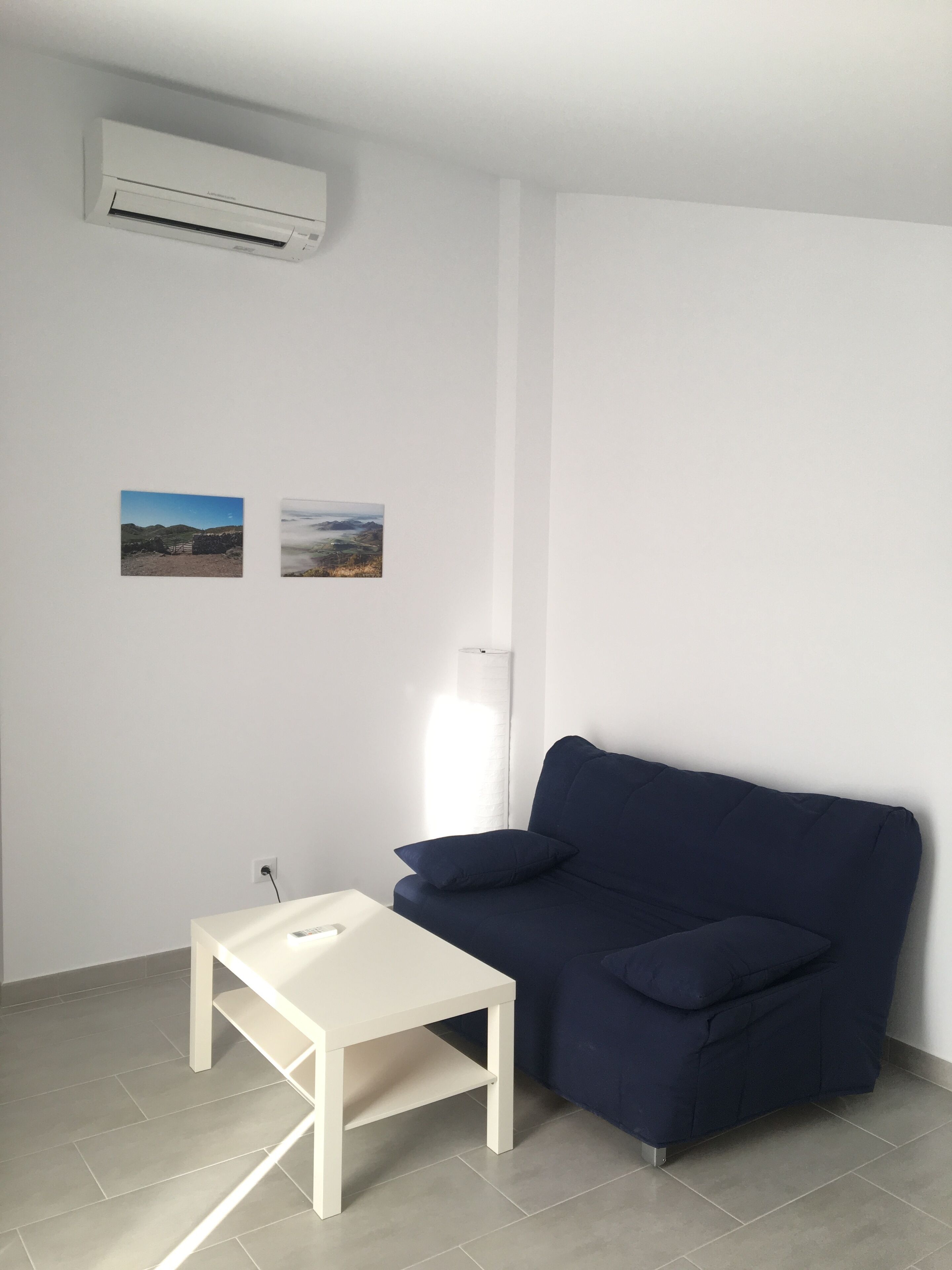 Una habitación con aire acondicionado blanco, una mesa blanca, un sofá azul oscuro y dos fotografías de paisajes en la pared.