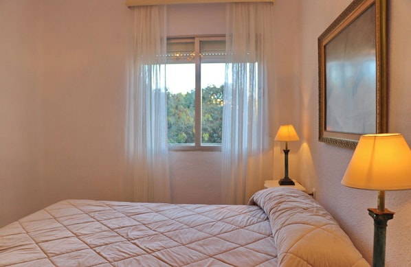 A hotel room with a bed, two bedside lamps, a window with sheer curtains, and a framed picture on the wall.