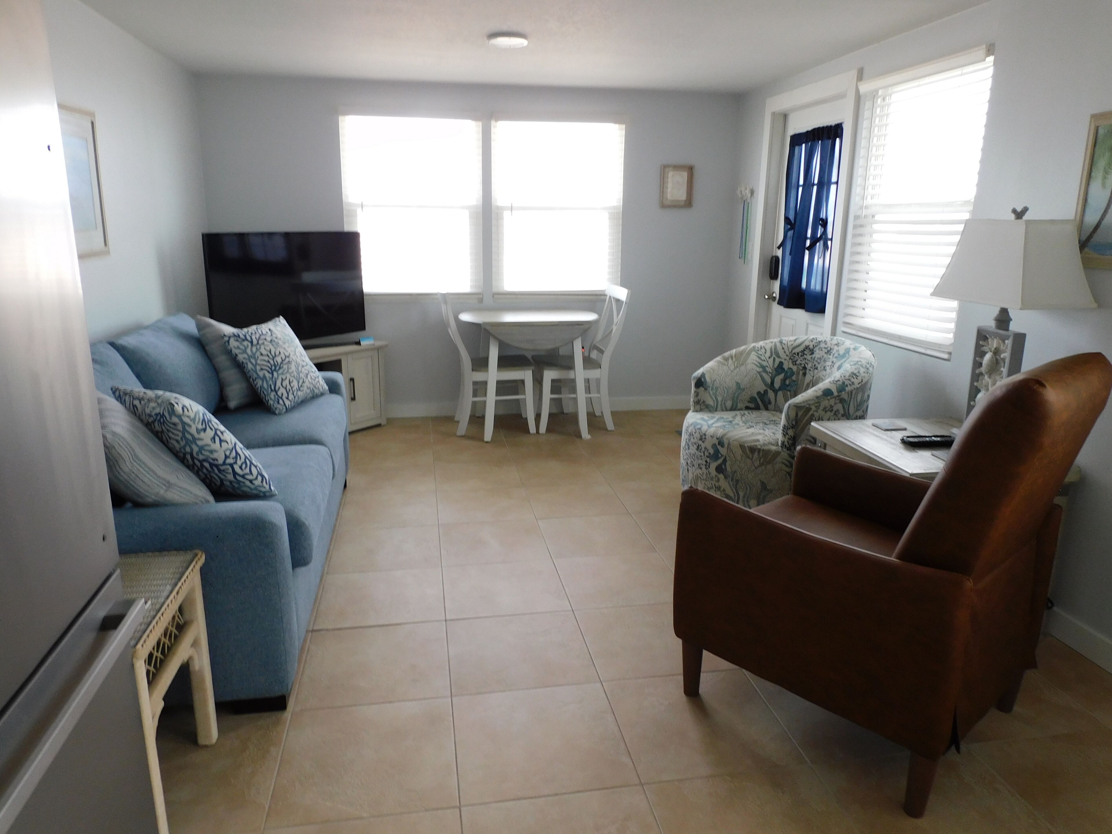 A living room with a sofa, a brown armchair, a TV, a dining table with chairs, and a window with blinds.