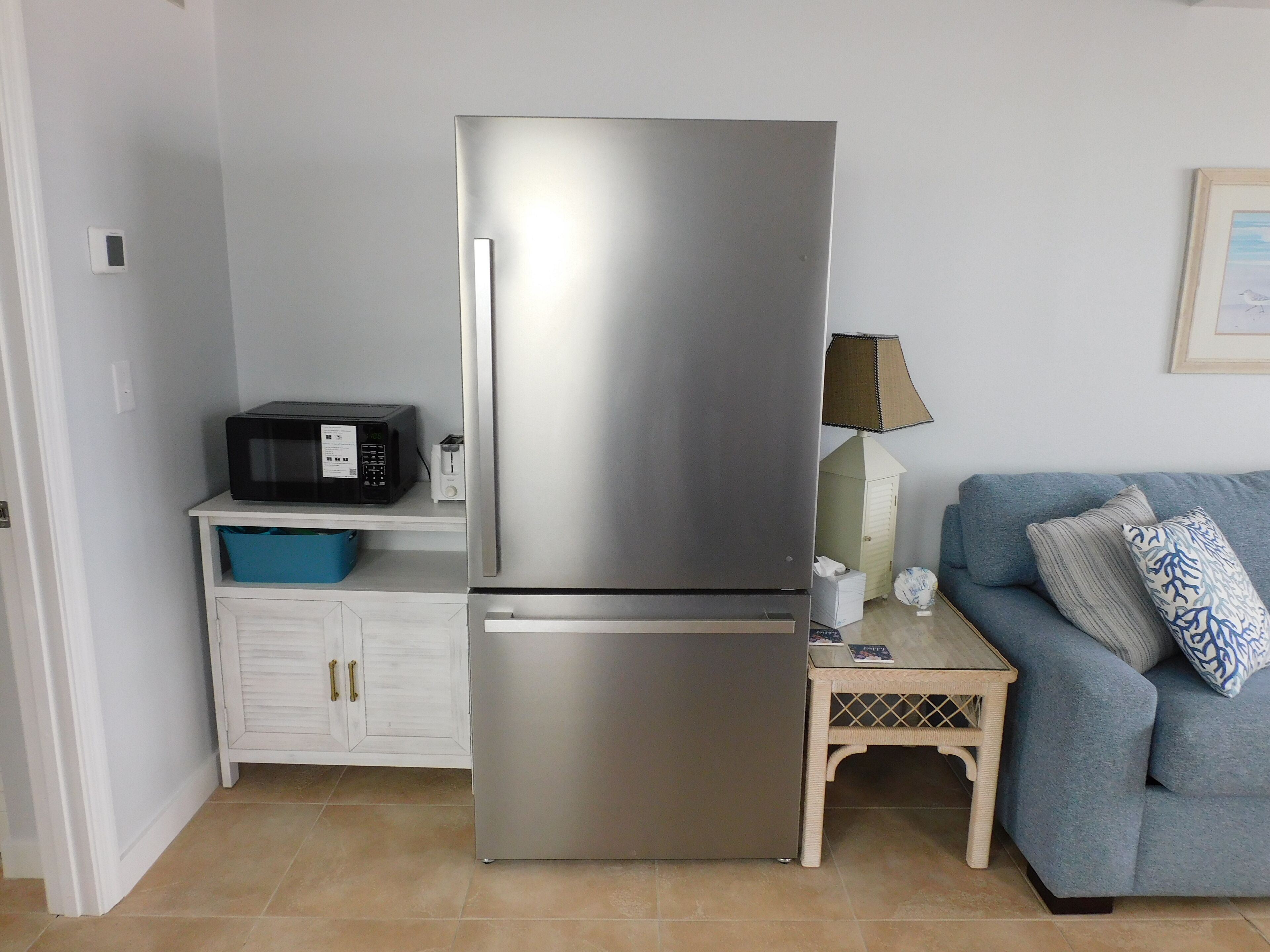A compact kitchen with a stainless steel refrigerator, a microwave, and a small table with a lamp.
