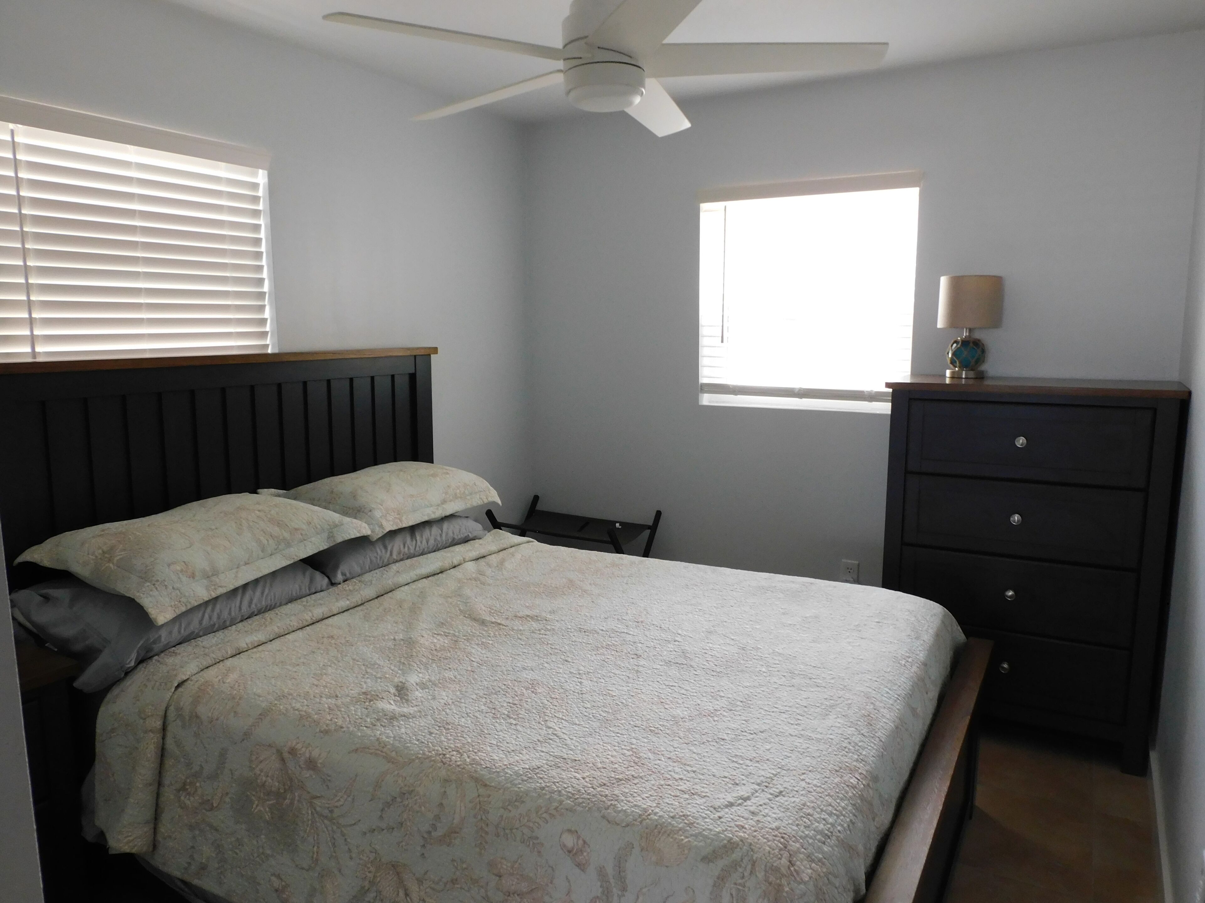 A bedroom with a bed, a dresser, a ceiling fan, a window, and a lamp.