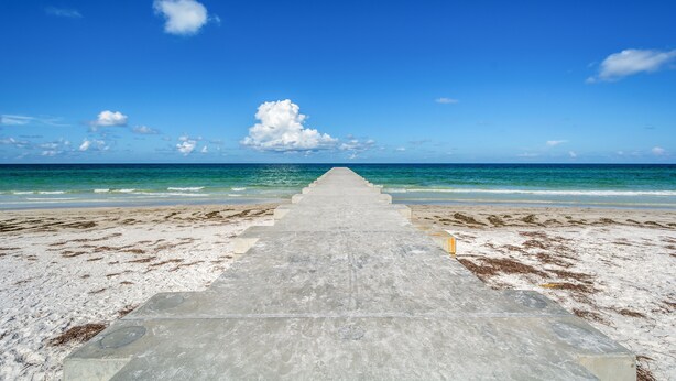 A concrete pathway leading to the beach with a clear blue sky and ocean in the background.
