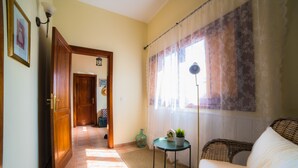 A living room with a wicker sofa, a small table with a plant, a lamp, and a view of a hallway with a wooden door. -  Peace in the countryside 1 km from the beach, magnificent views, bbq and free wifi (Tabayesco)