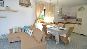 A living room with a sofa and chairs, a dining table set with plates and glasses, and a kitchen area with cabinets and a window. - BLUE BAY HOUSE (Marina San Gregorio)