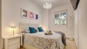 A bedroom with a bed, bedside table, lamp, artwork, and a window.